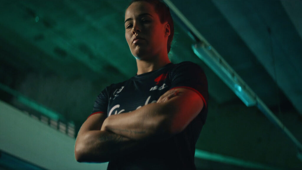An NRLW player standing with her arms folded in a stadium tunnel, lit in teal and red