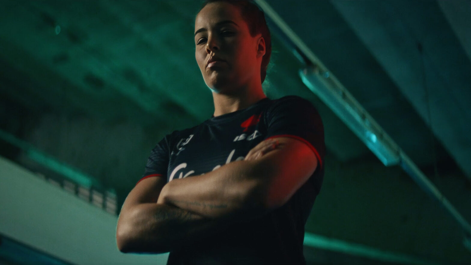 An NRLW player standing with her arms folded in a stadium tunnel, lit in teal and red